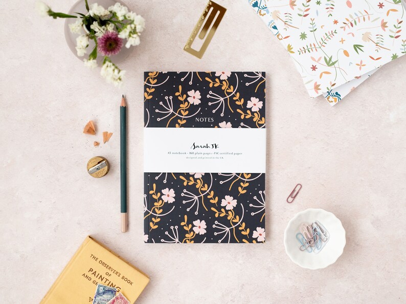 May include: A black notebook with a floral pattern in pink, yellow, and white. The notebook says "NOTES" and "Sarah JK" on the front cover. The notebook is on a light pink surface with a pencil, a pencil sharpener, and a book.