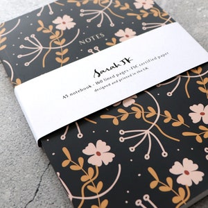 May include: A black A5 notebook with a floral pattern in shades of pink and gold. The notebook has 160 lined pages and is made with FSC certified paper. The cover has a white band with the text "NOTES" and "Sarah JK" in cursive. The band also includes the text "A5 notebook - 160 lined pages - FSC certified paper designed and printed in the UK."