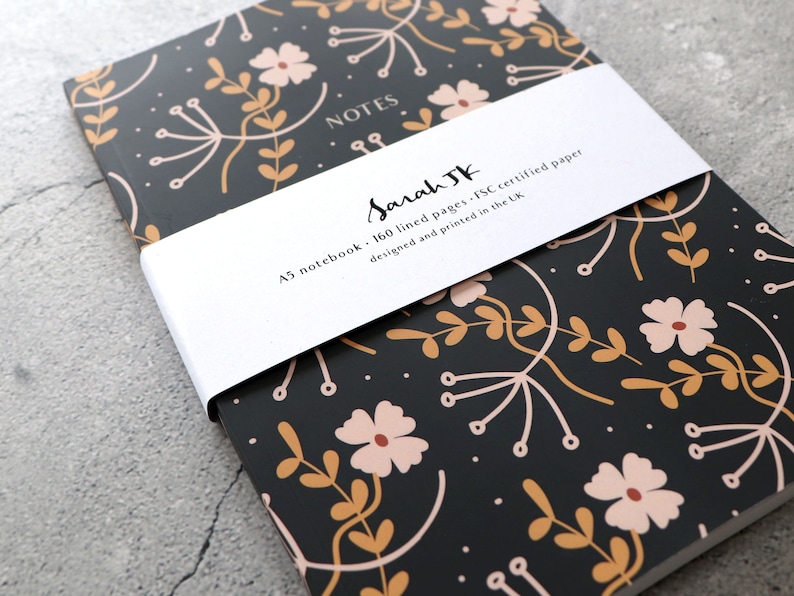 May include: A black A5 notebook with a floral pattern in shades of pink and gold. The notebook has 160 lined pages and is made with FSC certified paper. The cover has a white band with the text "NOTES" and "Sarah JK" in cursive. The band also includes the text "A5 notebook - 160 lined pages - FSC certified paper designed and printed in the UK."