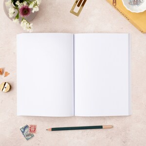 May include: An open blank notebook with a green pencil lying on a light beige surface. There are a few pencil shavings, a pencil sharpener, and some stamps near the notebook.