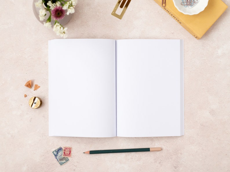 May include: An open blank notebook with a green pencil lying on a light beige surface. There are a few pencil shavings, a pencil sharpener, and some stamps near the notebook.
