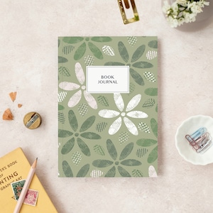 May include: A sage green book journal with a floral pattern of white and green flowers. The cover has a white rectangle with the text "BOOK JOURNAL". Stationery items, including a pencil and paperclips, are arranged around the journal.