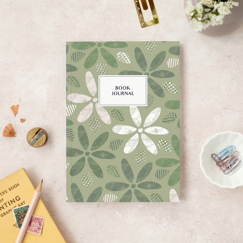 May include: A sage green book journal with a floral pattern of white and green flowers. The cover has a white rectangle with the text "BOOK JOURNAL". Stationery items, including a pencil and paperclips, are arranged around the journal.