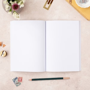 May include: An open blank notebook with a green pencil lying on a light beige surface. There are a few pencil shavings, a pencil sharpener, and some stamps near the notebook.