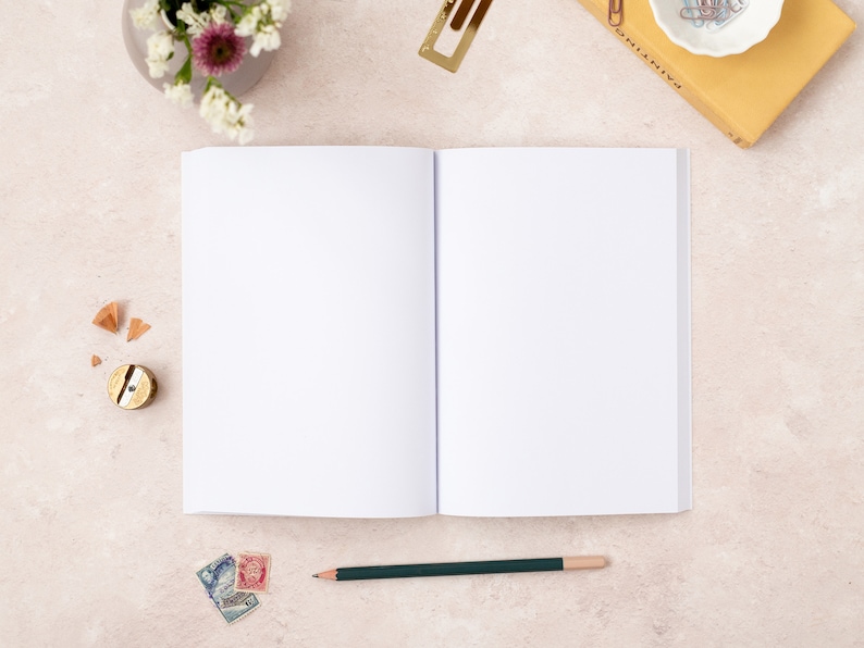 May include: An open blank notebook with a green pencil lying on a light beige surface. There are a few pencil shavings, a pencil sharpener, and some stamps near the notebook.