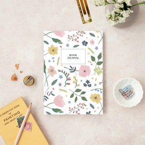 May include: A floral-patterned book journal with the text "BOOK JOURNAL" on the cover, surrounded by stationery and art supplies. The journal features a white background with pink, yellow, and blue flowers and green leaves. A yellow book and pencil are also visible.