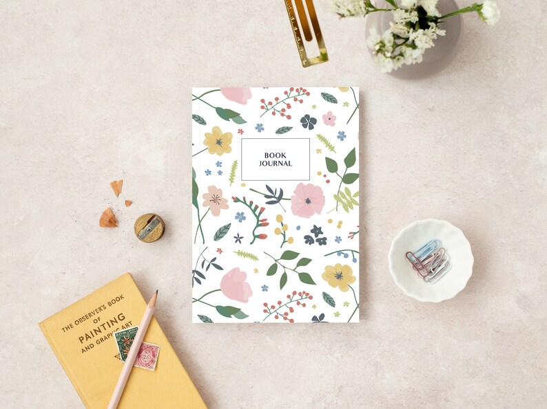May include: A floral-patterned book journal with the text "BOOK JOURNAL" on the cover, surrounded by stationery and art supplies. The journal features a white background with pink, yellow, and blue flowers and green leaves. A yellow book and pencil are also visible.