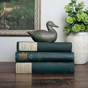 May include: A bronze-toned metal duck figurine resting on a stack of three dark green hardcover books. The top book is titled Saint Joan by Bernard Shaw, the middle is First in the Field by George Manville Fenn, and the bottom is American Nights Entertainment.
