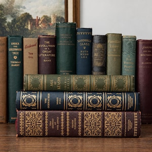 May include: A collection of vintage hardcover books arranged in a stack and a row. The books feature various colors including dark green, navy blue, brown, and maroon, with gold embossed lettering and decorative patterns on several of the spines.