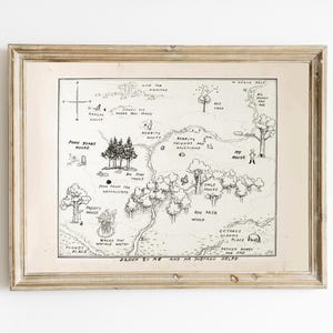 May include: A framed black and white map illustration of a whimsical forest with text. The map includes landmarks such as "Pooh Bear's House" and "Owl's House." The frame is a light brown colour.
