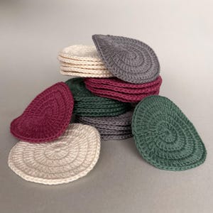 Puede incluir: Colección de discos desmaquillantes ganchillados en forma de lágrima, en crema, gris, burdeos y verde. Los discos están apilados y dispersos, mostrando su textura en espiral. Ideales para rutinas de cuidado de la piel.