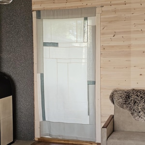 Pode incluir: Uma cortina de patchwork branca e cinza está pendurada em uma moldura de porta de madeira. A cortina é feita de painéis retangulares de vários tamanhos e tons de branco e cinza. A moldura da porta é de madeira clara.