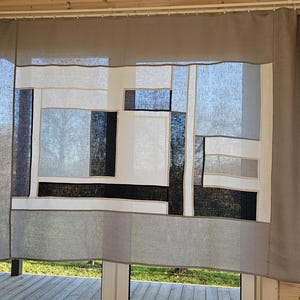 Può includere: Una tenda da finestra a motivi geometrici. La tenda presenta un design patchwork con forme rettangolari e quadrate in tonalità di grigio, nero, bianco e beige. La tenda è appesa davanti a una finestra con vista su un terrazzo in legno ed erba verde.