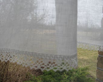May include: White sheer curtains with a lace trim, hanging in a window. The curtains are slightly translucent, allowing a view of the trees and foliage outside.