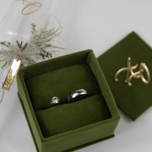 May include: A square olive green velvet ring box containing a silver-toned ring with a single round clear stone and a plain silver-toned band. The box lid features a gold-toned metal XO script accent, and a glass flute with gold text is nearby.
