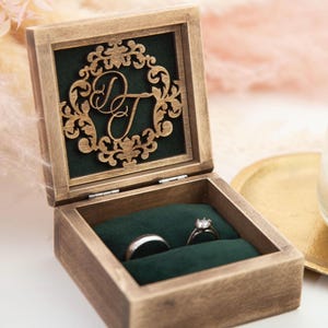 May include: Wooden ring box with a dark green velvet interior. The box is open, displaying two rings. The lid features a decorative wooden cutout with the initials "D T". The box is made of stained wood.