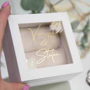 Può includere: Scatola per anelli bianca con coperchio trasparente e un'iscrizione dorata che recita "Vanessa & Stefan".