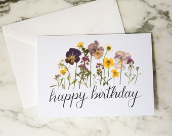 Op de afbeelding: Een witte kaart met een ontwerp van geperste bloemen en de tekst "happy birthday". De bloemen zijn paars, geel en wit.