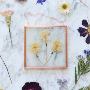 May include: A square, copper-framed glass piece with three dried yellow flowers. The piece hangs from a delicate chain. Scattered around are various dried flowers in shades of purple, white, and blue, set against a marble background.