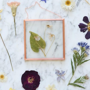 May include: A square, copper-framed glass display case with dried flowers and a green leaf. The case is suspended by a delicate chain. Various dried flowers in shades of purple, white, yellow, and blue are scattered around the frame.