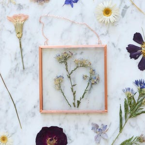 May include: A square copper-framed glass display with dried blue and white flowers. The display is hanging by a delicate chain. Various dried flowers in shades of purple, white, and yellow are scattered around the frame on a marble surface.