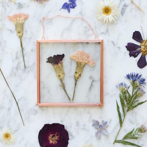 May include: A square, copper-colored framed pressed flower art piece with two dried flowers inside. The frame is suspended by a delicate chain. Surrounding the frame are various dried flowers in shades of purple, white, and yellow, scattered on a marble surface.