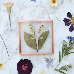 May include: A square, copper-framed glass display case holding pressed lily of the valley flowers and leaves. The case hangs from a delicate chain. Dried flowers, including daisies and purple blooms, are scattered on a marble surface.