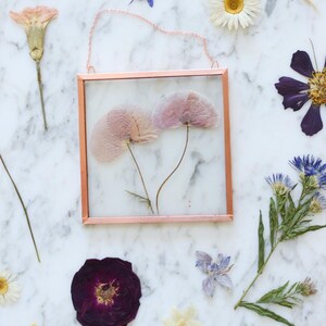 May include: A square, copper-colored framed pressed flower art piece with two pink flowers. The art is surrounded by various dried flowers in shades of purple, white, and yellow. A delicate chain is attached for hanging.