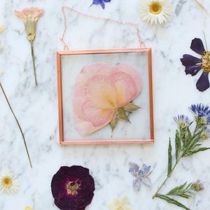 May include: A square, copper-colored framed pressed flower art piece with a pink rose. The art is surrounded by various dried flowers on a white marble surface. The frame has a delicate chain for hanging.