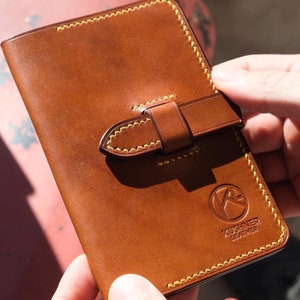 May include: A brown leather wallet with a yellow stitch detail and a strap closure. The wallet has a logo that says "Kushner Leather".