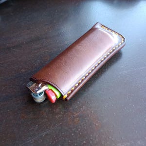 May include: A brown leather lighter case with yellow stitching. The case holds a silver lighter with red and green accents. The lighter case is on a dark brown surface.
