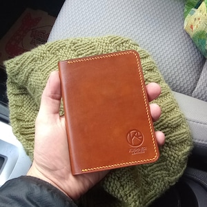 Puede incluir: Una cartera de cuero marrón con detalles de costura amarilla y el texto "Kushner Leather" grabado en la parte delantera.