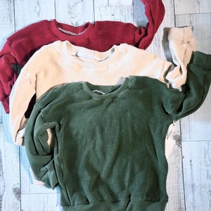 May include: Three sweaters stacked on top of each other. The top sweater is red, the middle sweater is cream, and the bottom sweater is green.