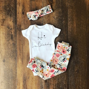 Pu&ograve; includere: Un completo per neonati composto da un body bianco con la scritta "Hi I'm Blakely" in nero, abbinato a leggings con stampa floreale e una fascia coordinata. Il motivo floreale presenta rose rosa, foglie verdi e dettagli dorati su sfondo crema.
