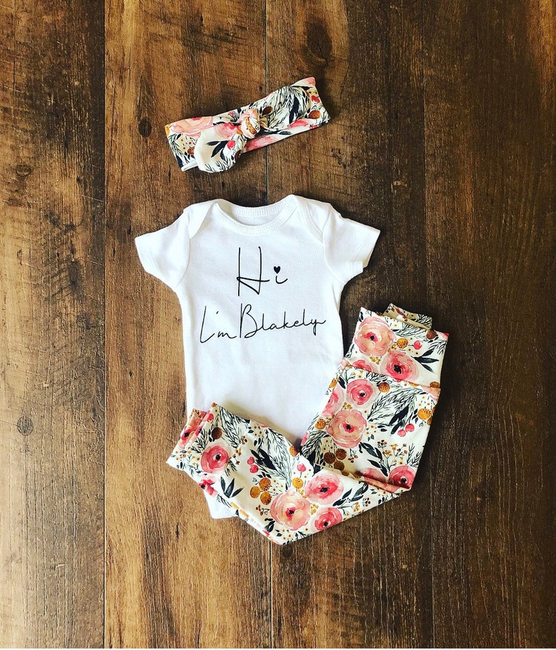 Pu&ograve; includere: Un completo per neonati composto da un body bianco con la scritta "Hi I'm Blakely" in nero, abbinato a leggings con stampa floreale e una fascia coordinata. Il motivo floreale presenta rose rosa, foglie verdi e dettagli dorati su sfondo crema.