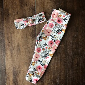 Pu&ograve; includere: Un set coordinato di leggings e fascia per neonati. Gli articoli sono bianchi con un motivo floreale di rose rosa, foglie nere e dettagli dorati. La fascia &egrave; annodata nella parte superiore.