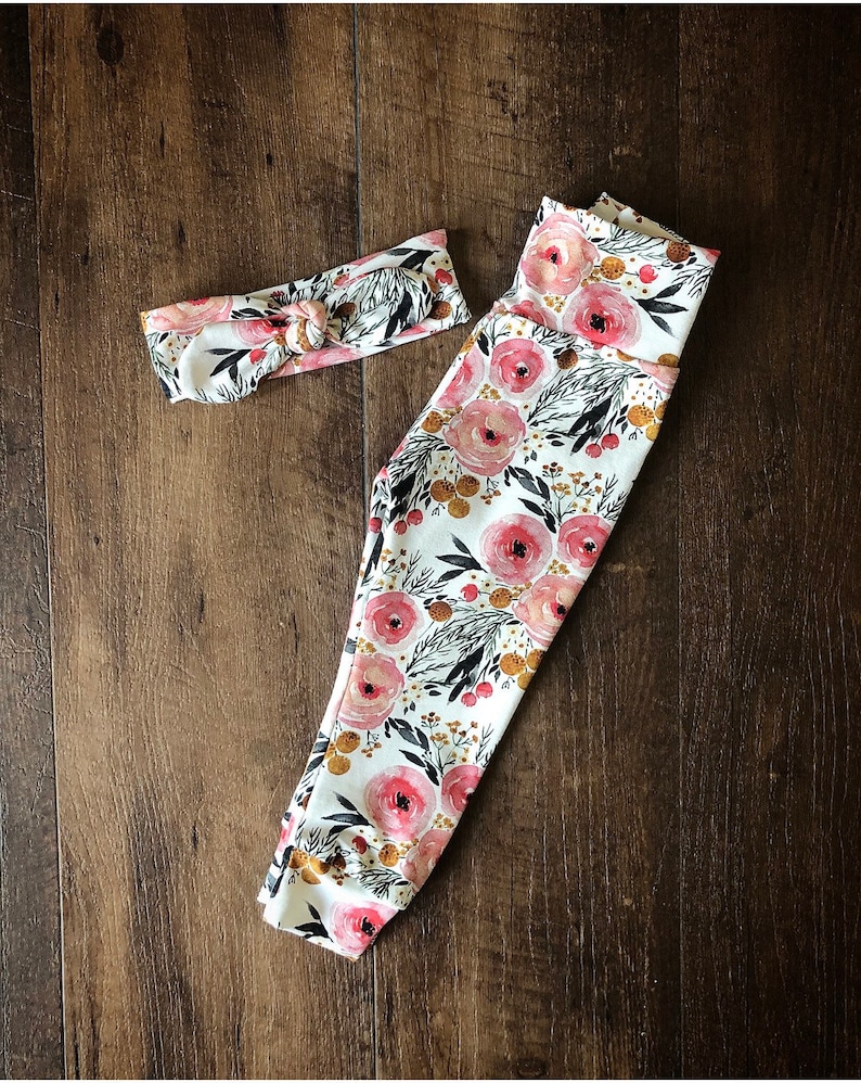 Pu&ograve; includere: Un set coordinato di leggings e fascia per neonati. Gli articoli sono bianchi con un motivo floreale di rose rosa, foglie nere e dettagli dorati. La fascia &egrave; annodata nella parte superiore.