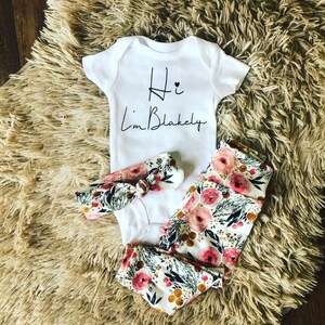 Pu&ograve; includere: Un body bianco per neonati con la scritta "Hi I'm Blakely" in corsivo nero, abbinato a leggings con stampa floreale, una fascia abbinata e un bavaglino. Il motivo floreale presenta rose rosa, foglie verdi e dettagli dorati.