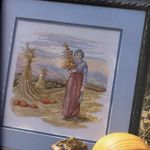 May include: Cross-stitch artwork in a framed presentation. The scene depicts a person in a long skirt and bonnet holding a bundle of gourds in a field with pumpkins and haystacks. The sky is a mix of blue, orange, and gray.