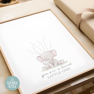 May include: A framed print with a watercolor illustration of a gray elephant with pink ears. The elephant is sitting on a green patch of grass. The text "you are so loved little one" is written in gray below the elephant.