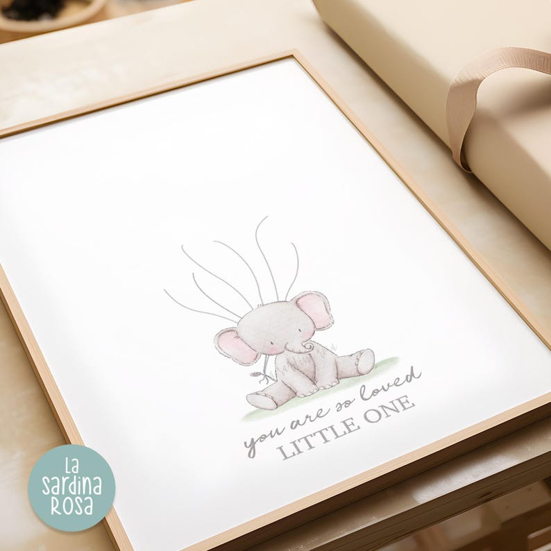 May include: A framed print with a watercolor illustration of a gray elephant with pink ears. The elephant is sitting on a green patch of grass. The text "you are so loved little one" is written in gray below the elephant.