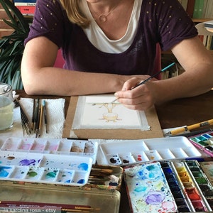 Puede incluir: Una persona pinta una ilustraci&oacute;n de acuarela de una jirafa en una hoja de papel blanca. El artista est&aacute; usando un pincel peque&ntilde;o y una paleta de acuarelas. La ilustraci&oacute;n est&aacute; en una tabla de madera.