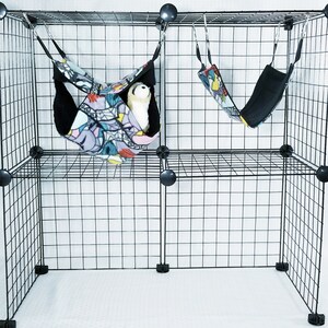May include: A black wire cage with two hanging pet hammocks. One hammock contains a white stuffed animal. The hammocks have a colorful geometric pattern on the outside and black fleece on the inside.