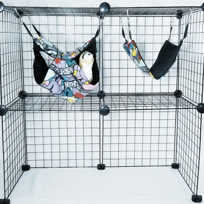 May include: A black wire cage with two hanging pet hammocks. One hammock contains a white stuffed animal. The hammocks have a colorful geometric pattern on the outside and black fleece on the inside.