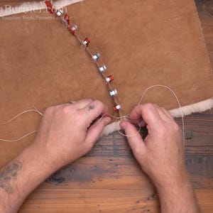 May include: A close-up of leatherwork in progress. A person's hands are shown stitching together two pieces of brown leather with white thread. The leather is held together with metal clips.