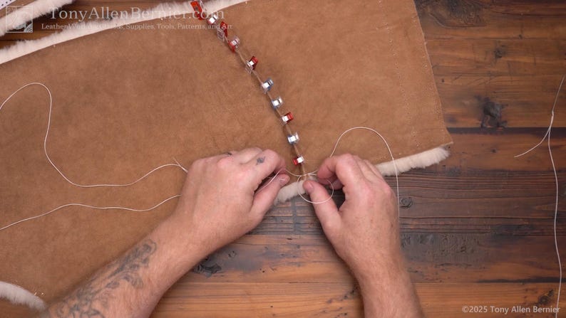 May include: A close-up of leatherwork in progress. A person's hands are shown stitching together two pieces of brown leather with white thread. The leather is held together with metal clips.