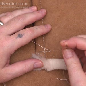 May include: Close-up of hands sewing a light brown leather material with white thread. The needle is being pushed through the leather, which has a white, fluffy trim. The image shows a detailed view of the stitching process.