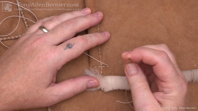 May include: Close-up of hands sewing a light brown leather material with white thread. The needle is being pushed through the leather, which has a white, fluffy trim. The image shows a detailed view of the stitching process.