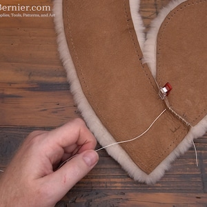 May include: A person is sewing two pieces of brown leather together with white thread. The leather is folded over and the edges are being stitched together. A red clamp is holding the leather in place.