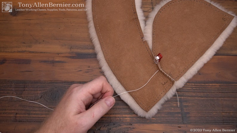 May include: A person is sewing two pieces of brown leather together with white thread. The leather is folded over and the edges are being stitched together. A red clamp is holding the leather in place.
